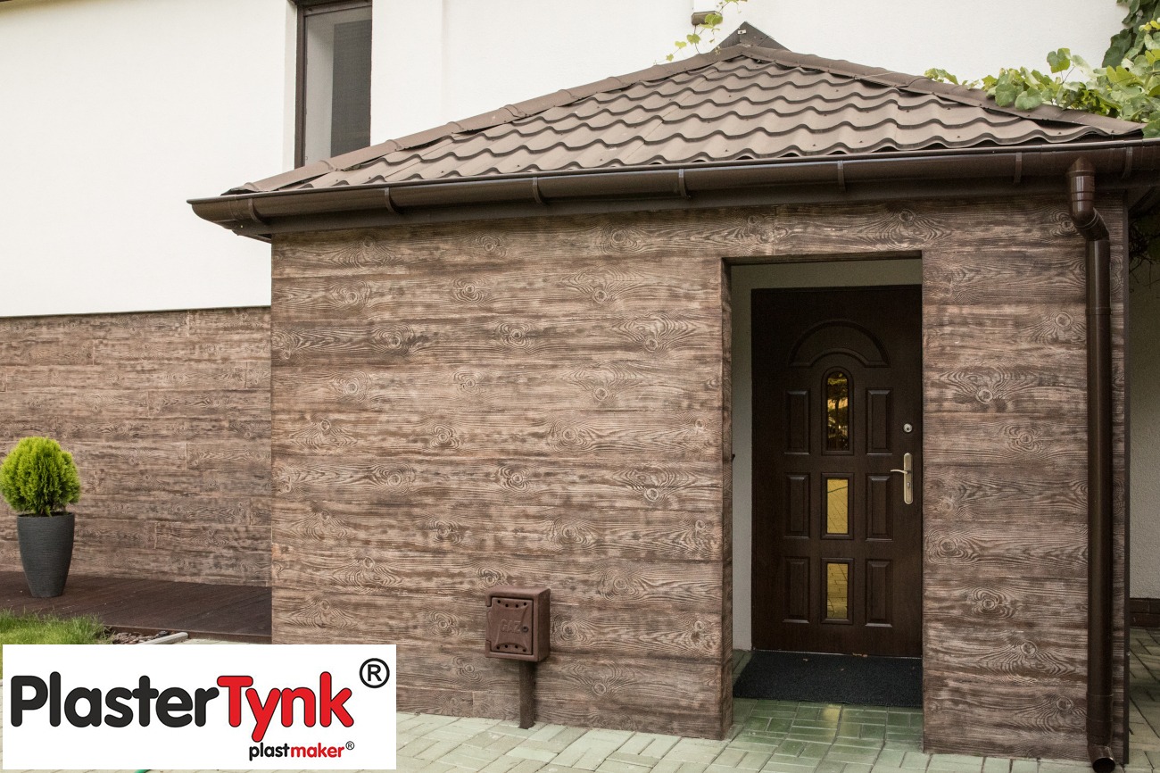 Zadaszone wejście do budynku wykończone panelami elewacyjnymi imitującymi drewno, z widoczną brązową stolarką drzwiową i małą skrzynką pocztową przy ścianie. W tle fragment białej ściany budynku...