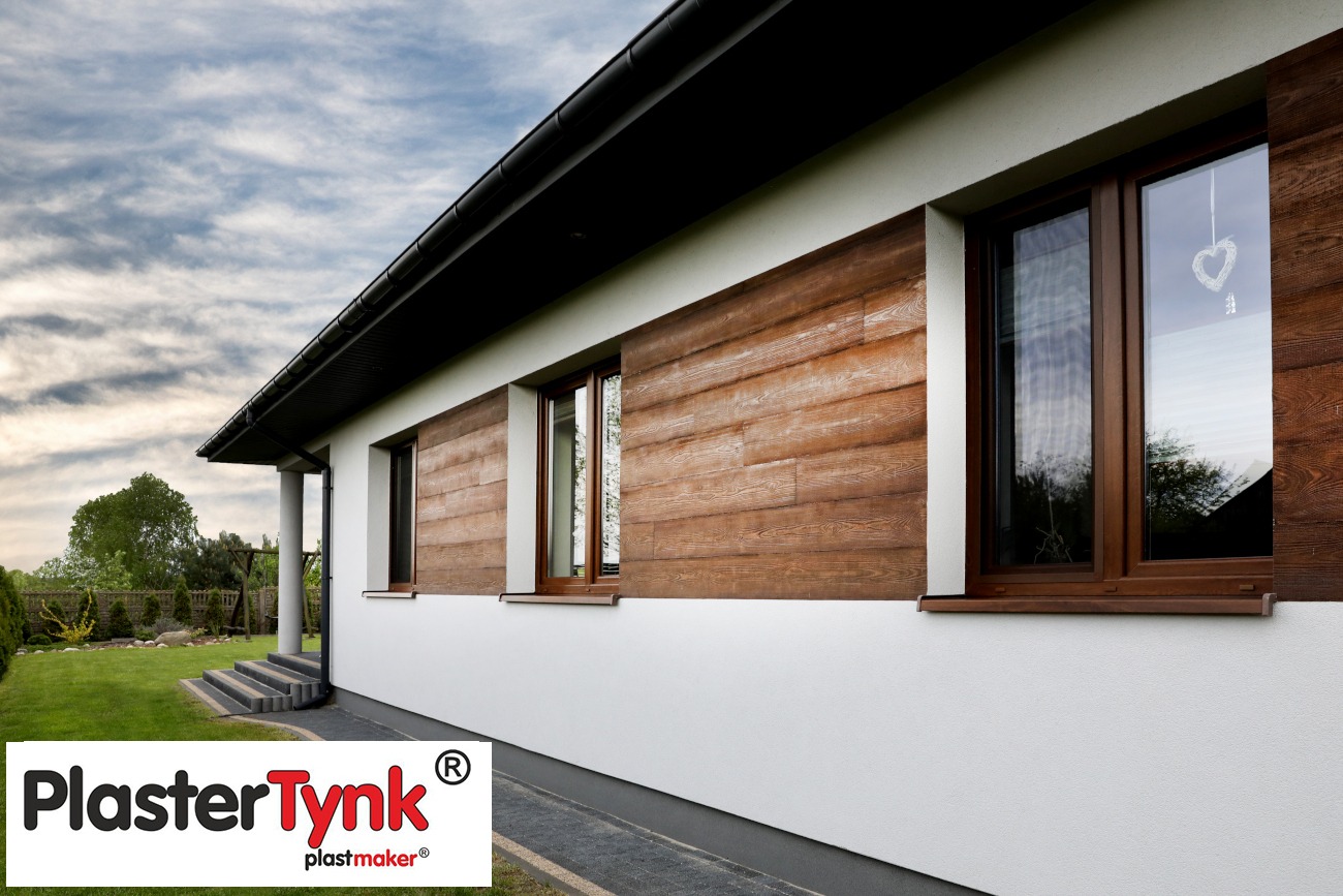 Dom jednorodzinny z elewacją imitującą drewno, biały tynk na dole, brązowe ramy okienne, w oknie zawieszka w kształcie serca, widoczny ogród i logo producenta tynku.