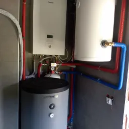 Zestaw urządzeń grzewczych: pompa ciepła Panasonic Aquarea, dwa zbiorniki akumulacyjne (jeden szary marki Trinnity), orurowanie w izolacji czerwonej i niebieskiej, zamontowane na tle szarych płytek.