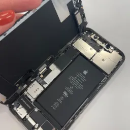 Otwarty iPhone z widoczną baterią, elementami wewnętrznymi i częściowo odpiętym ekranem, trzymanym przez dłoń z czerwonym lakierem do paznokci.