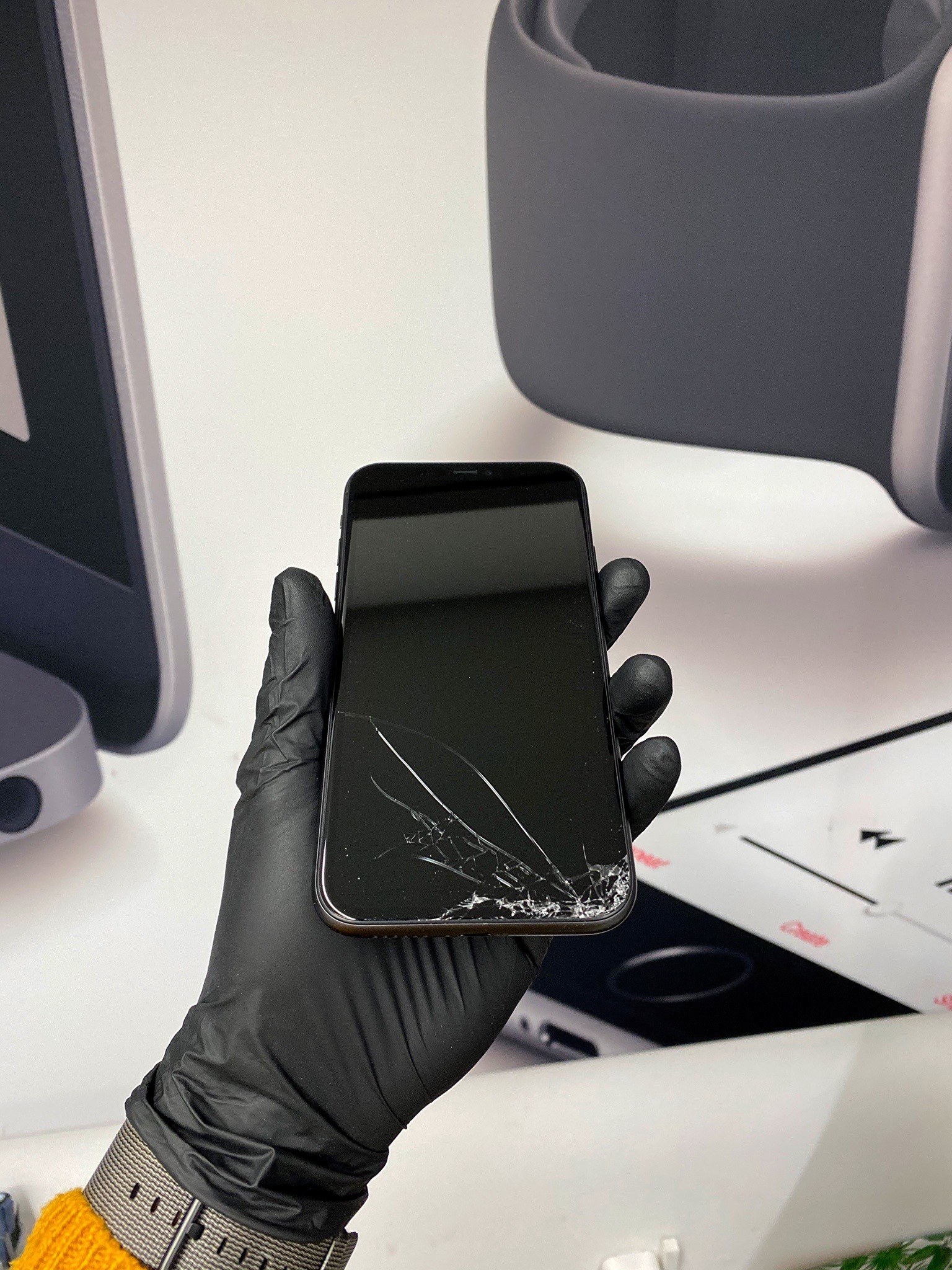 Smartfon z pękniętym ekranem trzymany w dłoni ubranej w czarną lateksową rękawiczkę, na tle grafiki przedstawiającej monitor i smartwatch.