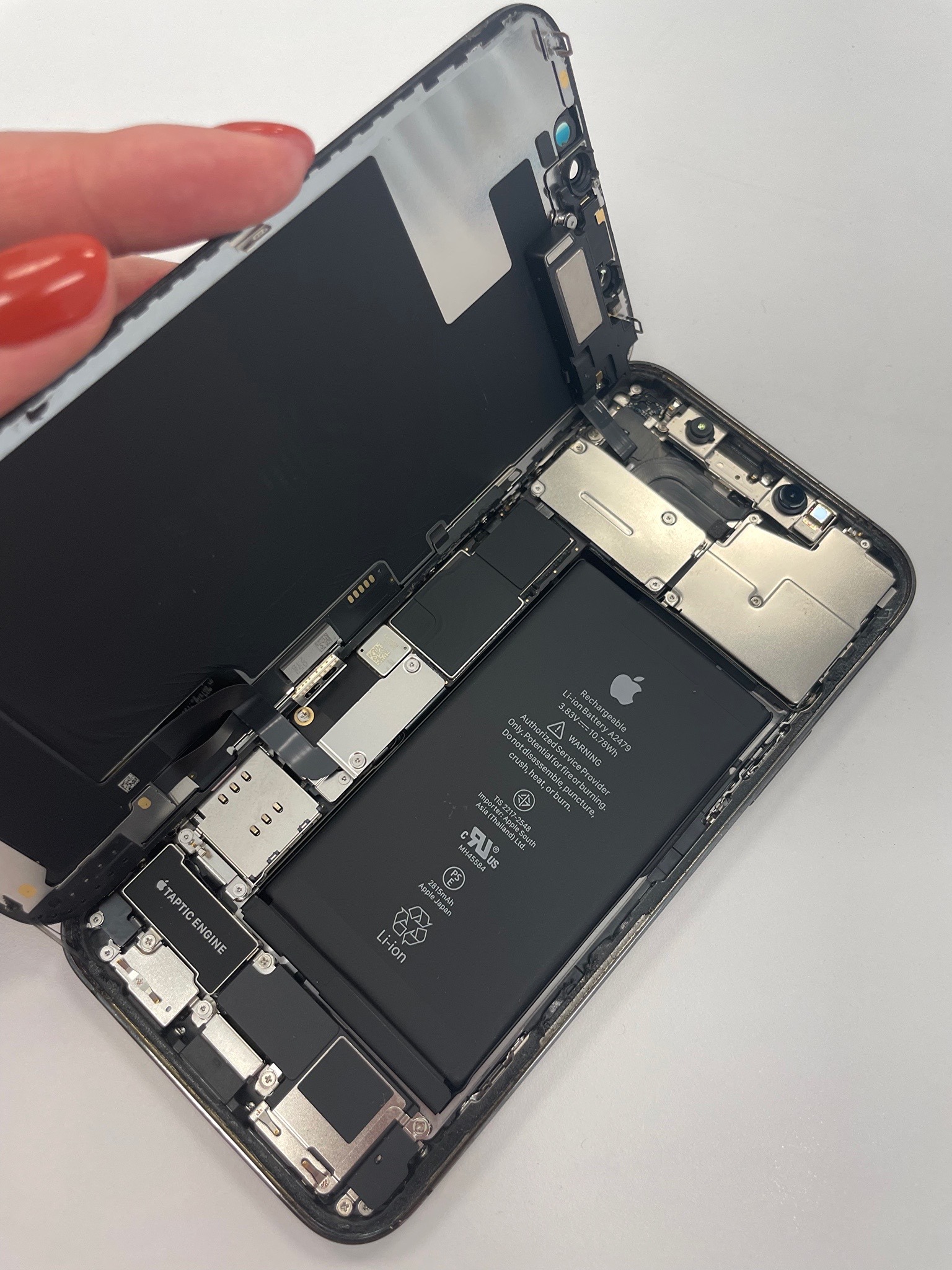 Otwarty iPhone z widoczną baterią, elementami wewnętrznymi i częściowo odpiętym ekranem, trzymanym przez dłoń z czerwonym lakierem do paznokci.