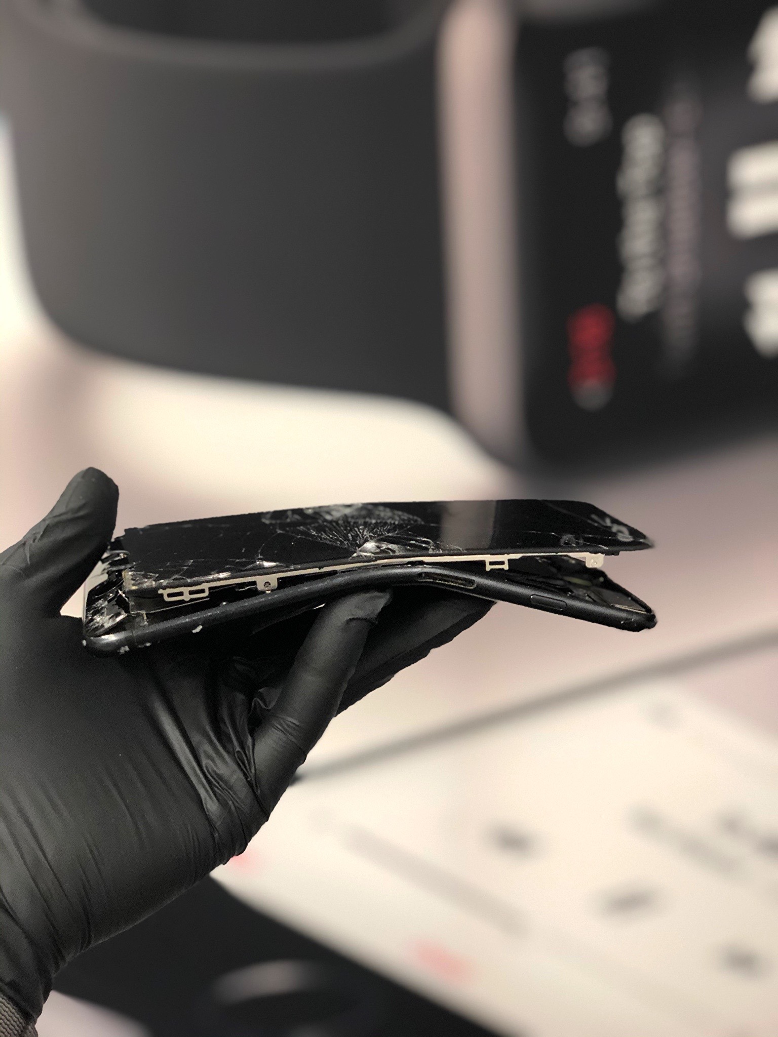 Smartfon z rozbitym ekranem, trzymany w dłoni w czarnej rękawiczce ochronnej, widoczne uszkodzenia mechaniczne obudowy i wyświetlacza.