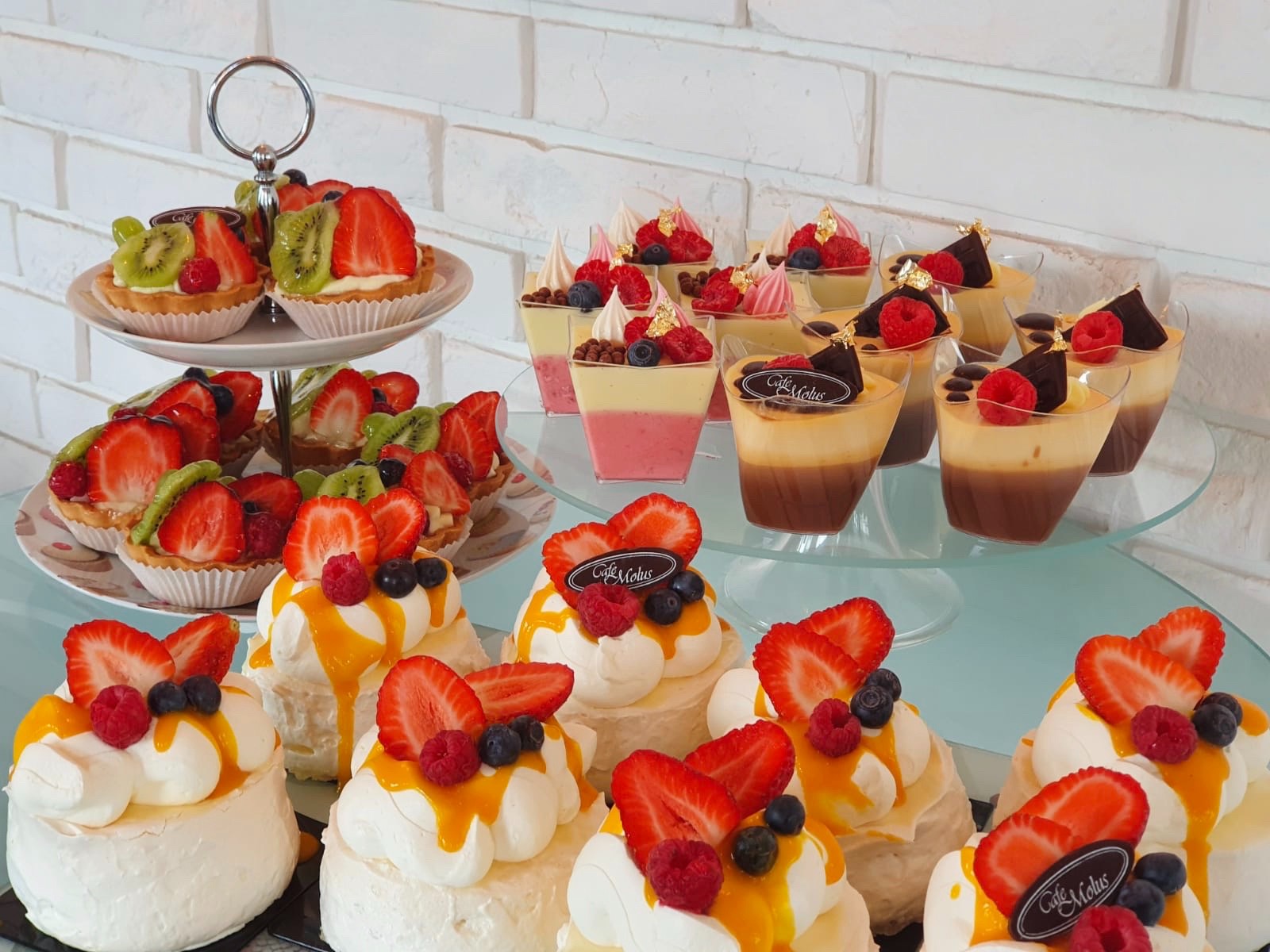 Elegancka ekspozycja deserów: mini bezy z sosem mango i owocami, tartaletki z kiwi i truskawkami na dwupoziomowym stojaku oraz warstwowe desery w kubeczkach z owocami i złotymi płatkami, na tle...