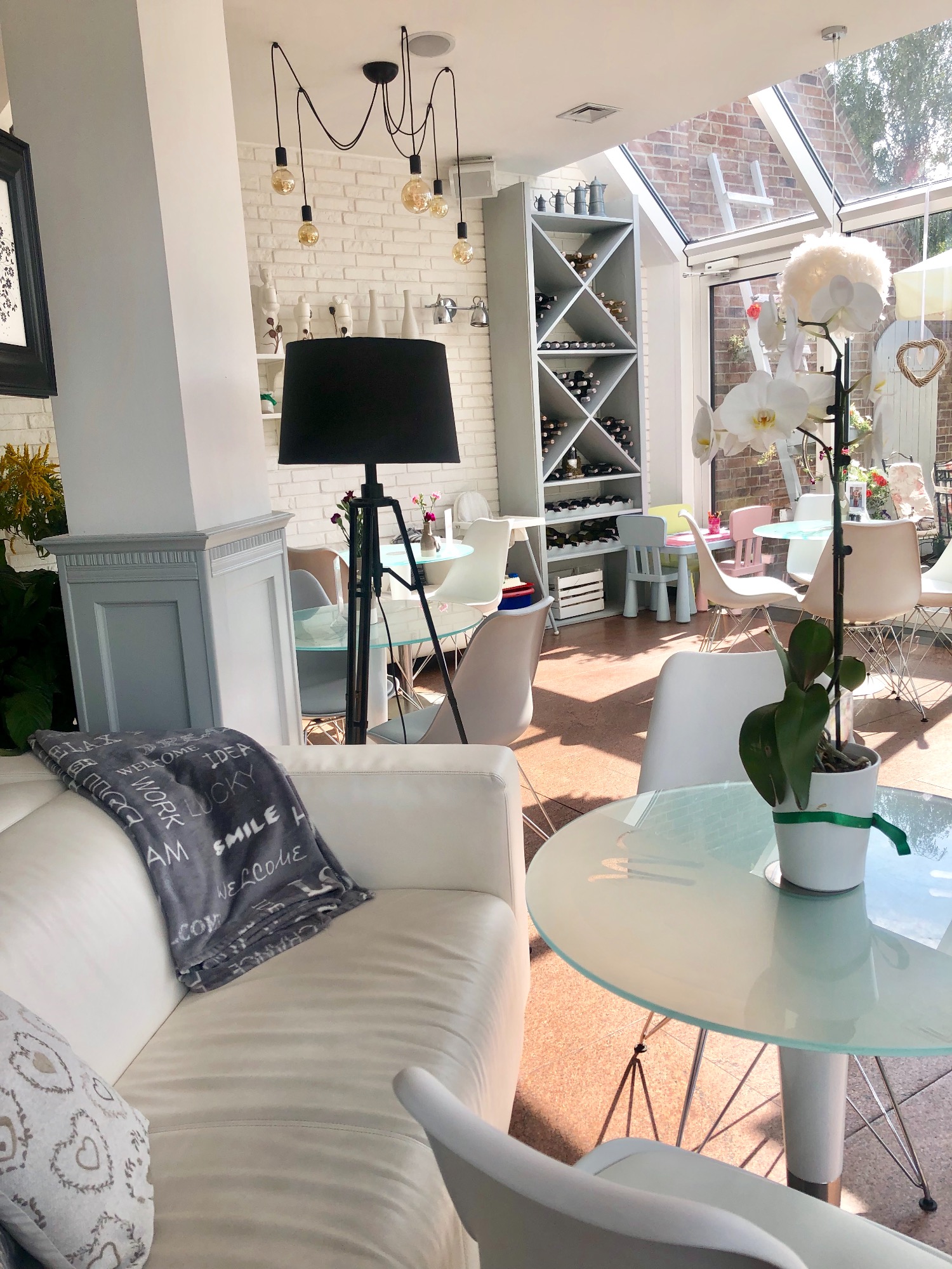 Jasne wnętrze kawiarni z białymi meblami, szklanymi stolikami, dekoracyjną lampą wiszącą i regałem na wino w tle, doświetlone naturalnym światłem przez przeszklony dach.