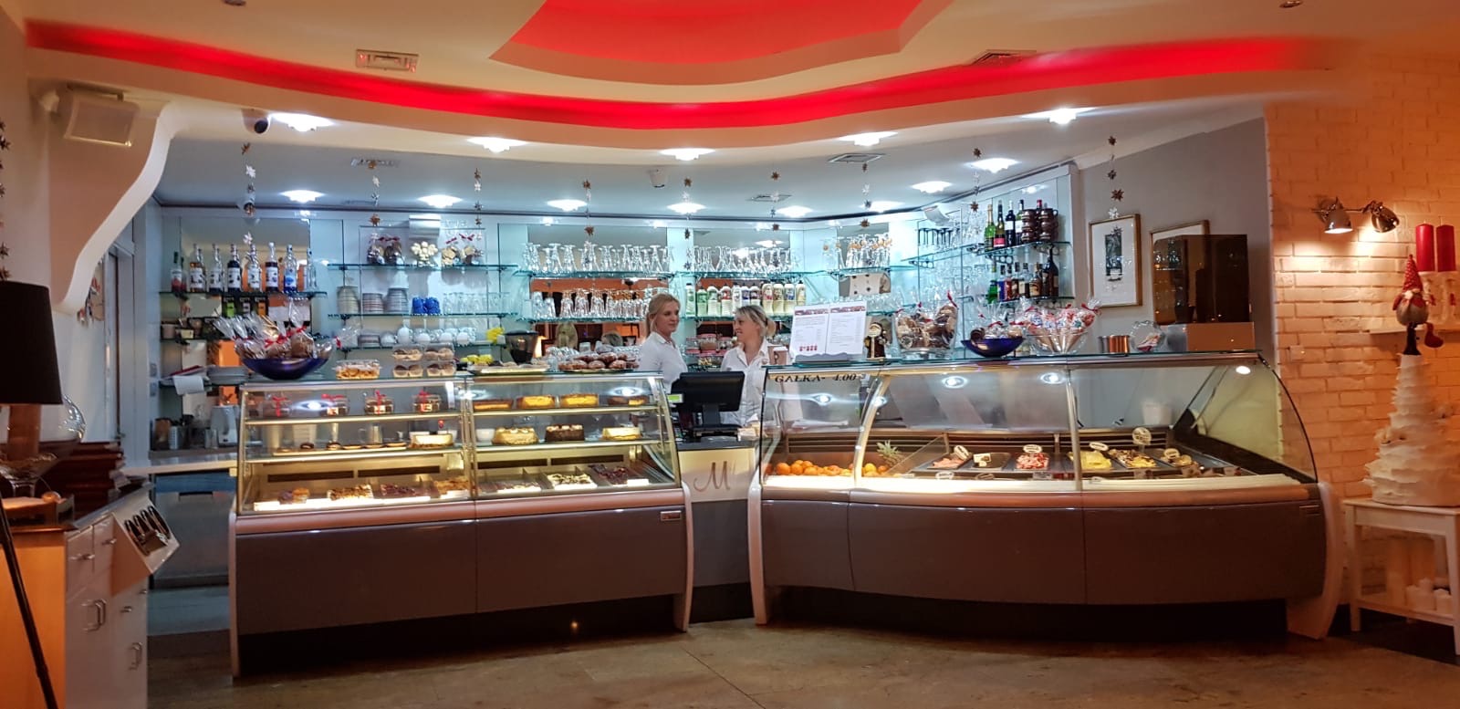 Wnętrze lokalu z witrynami pełnymi ciast i deserów, dwie kobiety w białych koszulach za ladą, półki z napojami i dekoracjami w tle, czerwone oświetlenie sufitowe.