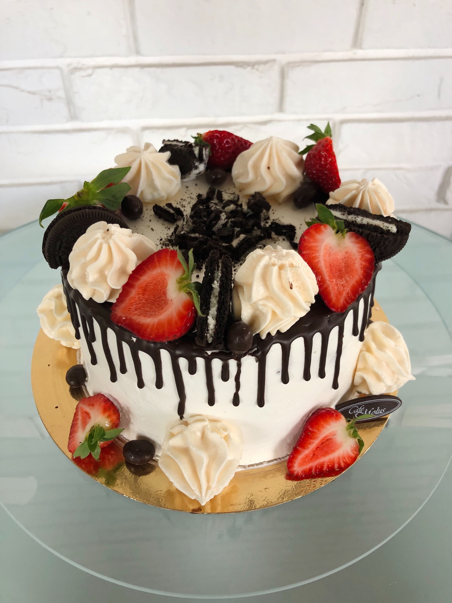 Tort udekorowany polewą czekoladową, truskawkami, bezami i pokruszonymi ciastkami Oreo, umieszczony na złotej podkładce na szklanym stole.