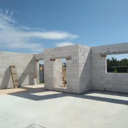 Wnętrze budynku w stanie surowym otwartym, ściany z białych bloczków, widoczne otwory okienne i drzwiowe, w tle błękitne niebo z chmurami i przenośna toaleta.