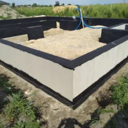 Wykop pod fundamenty z izolacją przeciwwilgociową, widoczne fragmenty zbrojenia i niebieskie rury instalacyjne.