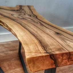 Perroc - Stolik kawowy z blatem z litego drewna orzechowego o nieregularnym kształcie, wsparty na metalowej, czarnej ramie, widoczny na tle podłogi imitującej drewno i białej ściany z gniazdkiem elektrycznym.