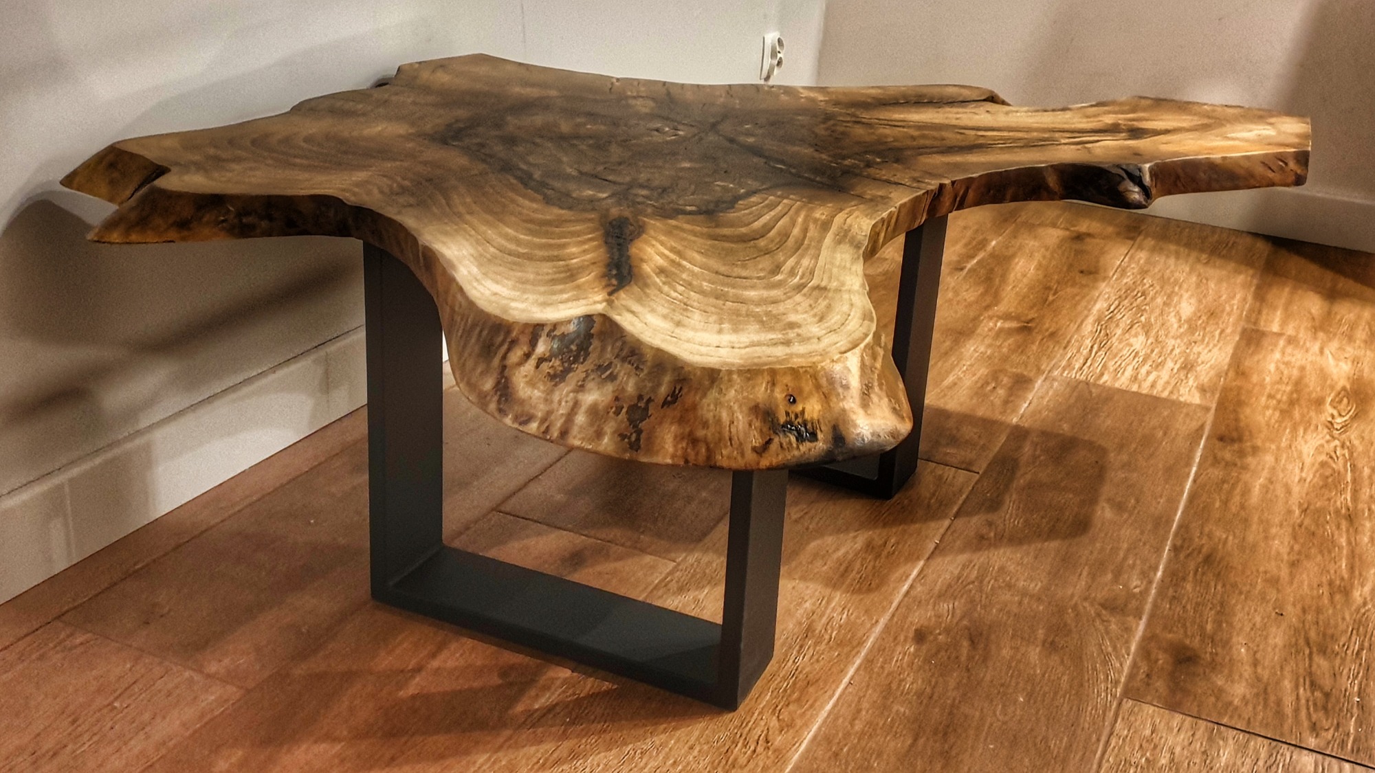 Stolik kawowy z blatem z naturalnego drewna o nieregularnym kształcie, osadzony na metalowych, czarnych nogach o kwadratowym przekroju, na tle drewnianej podłogi.