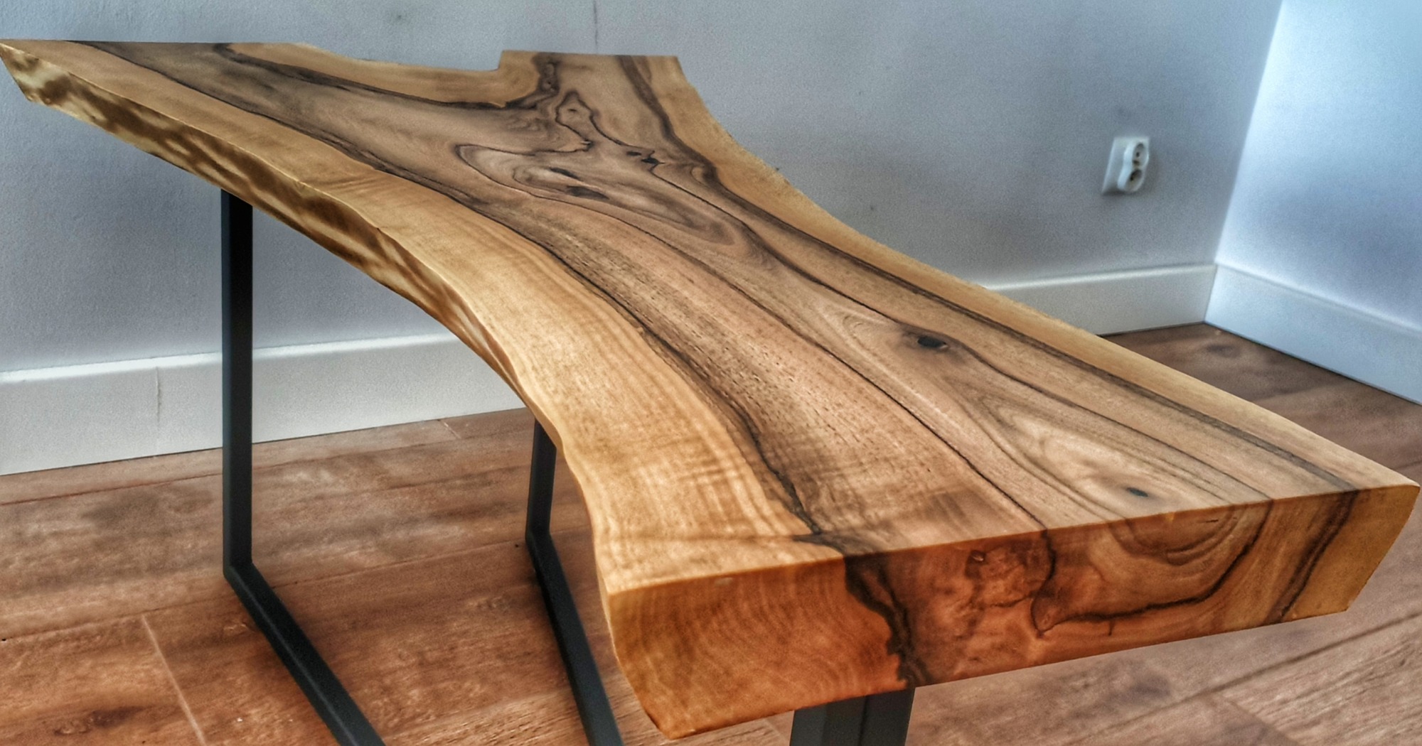 Stolik kawowy z blatem z litego drewna orzechowego o nieregularnym kształcie, wsparty na metalowej, czarnej ramie, widoczny na tle podłogi imitującej drewno i białej ściany z gniazdkiem elektrycznym.