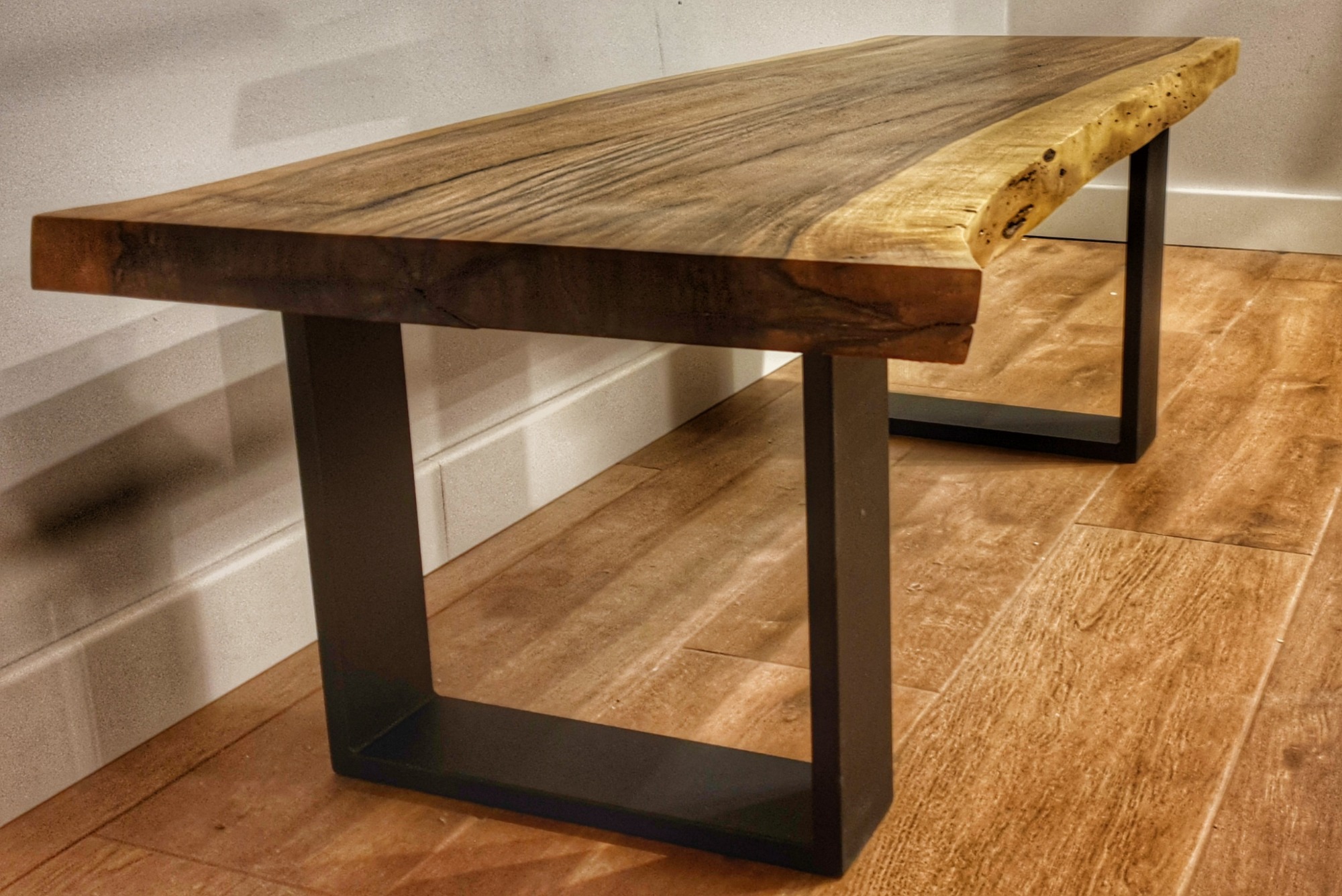 Ławka z blatem z litego drewna o nieregularnej krawędzi i ciemnych, metalowych nogach w kształcie prostokątów, na podłodze z paneli.