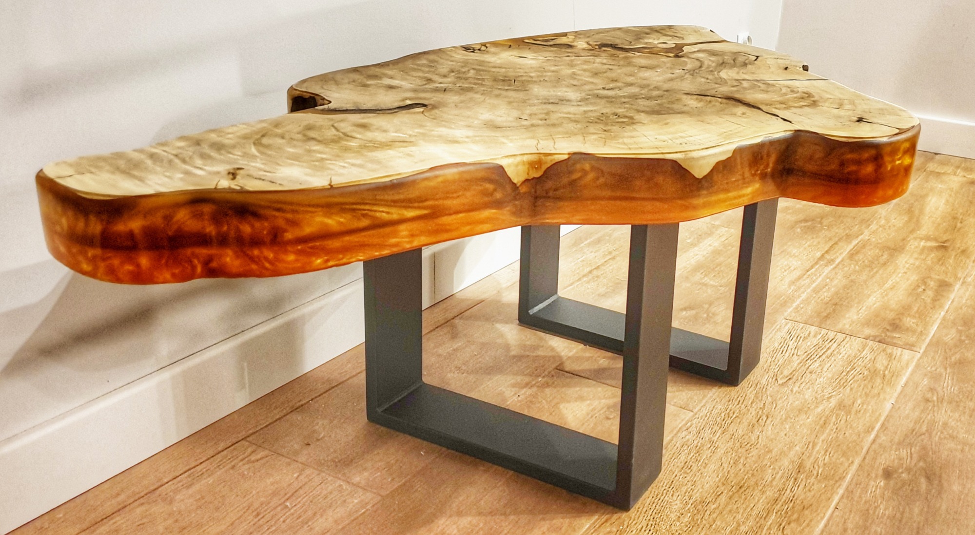 Stolik kawowy z blatem z litego drewna o nieregularnym kształcie, pokryty żywicą epoksydową w odcieniach bursztynu, na metalowych nogach w kształcie prostokątów.