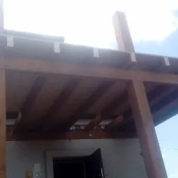 Drewniana konstrukcja szkieletowa budynku w trakcie budowy, widok z dołu na drewniane belki i słupy, częściowo zadaszona płytami, z fragmentem dachu istniejącego budynku w tle.