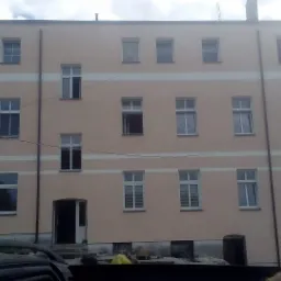 Trzypiętrowy budynek po renowacji elewacji, z beżową fasadą i białymi obramowaniami okien oraz poziomymi pasami. Widoczne okna z białymi ramami i częściowo zasłoniętymi roletami. Część budynku...
