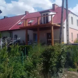 Drewniany balkon dobudowany do domu z czerwoną dachówką, widok z poziomu gruntu, częściowo zasłonięty przez zieleń.