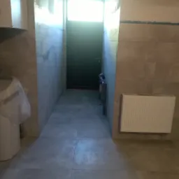 Wnętrze łazienki w trakcie remontu z szarymi płytkami podłogowymi i ściennymi, widoczne grzejniki, instalacja wodna i zakryta folią toaleta.