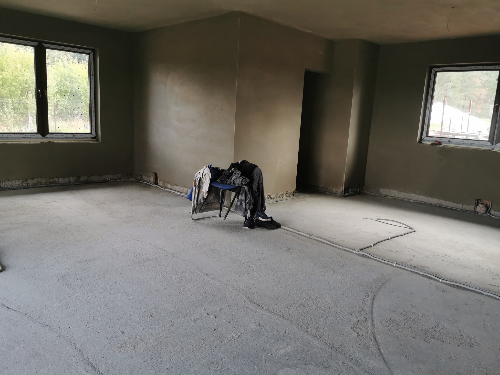 Pokój po nałożeniu szarego tynku maszynowego na ściany; na podłodze widoczne przewody elektryczne, a na krześle leżą ubrania robocze.