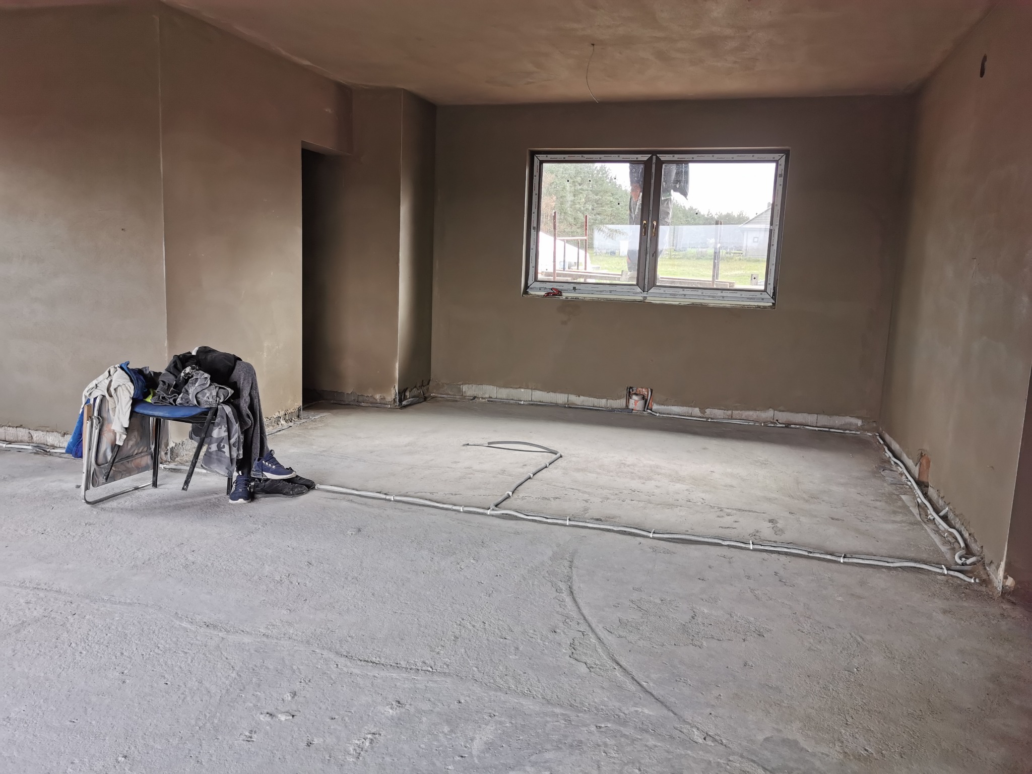 Pokój po nałożeniu tynków maszynowych, widoczne ściany w kolorze beżowym, okno z widokiem na zewnątrz, krzesło z ubraniami i butami w rogu pomieszczenia oraz przewody elektryczne ułożone na podłodze.