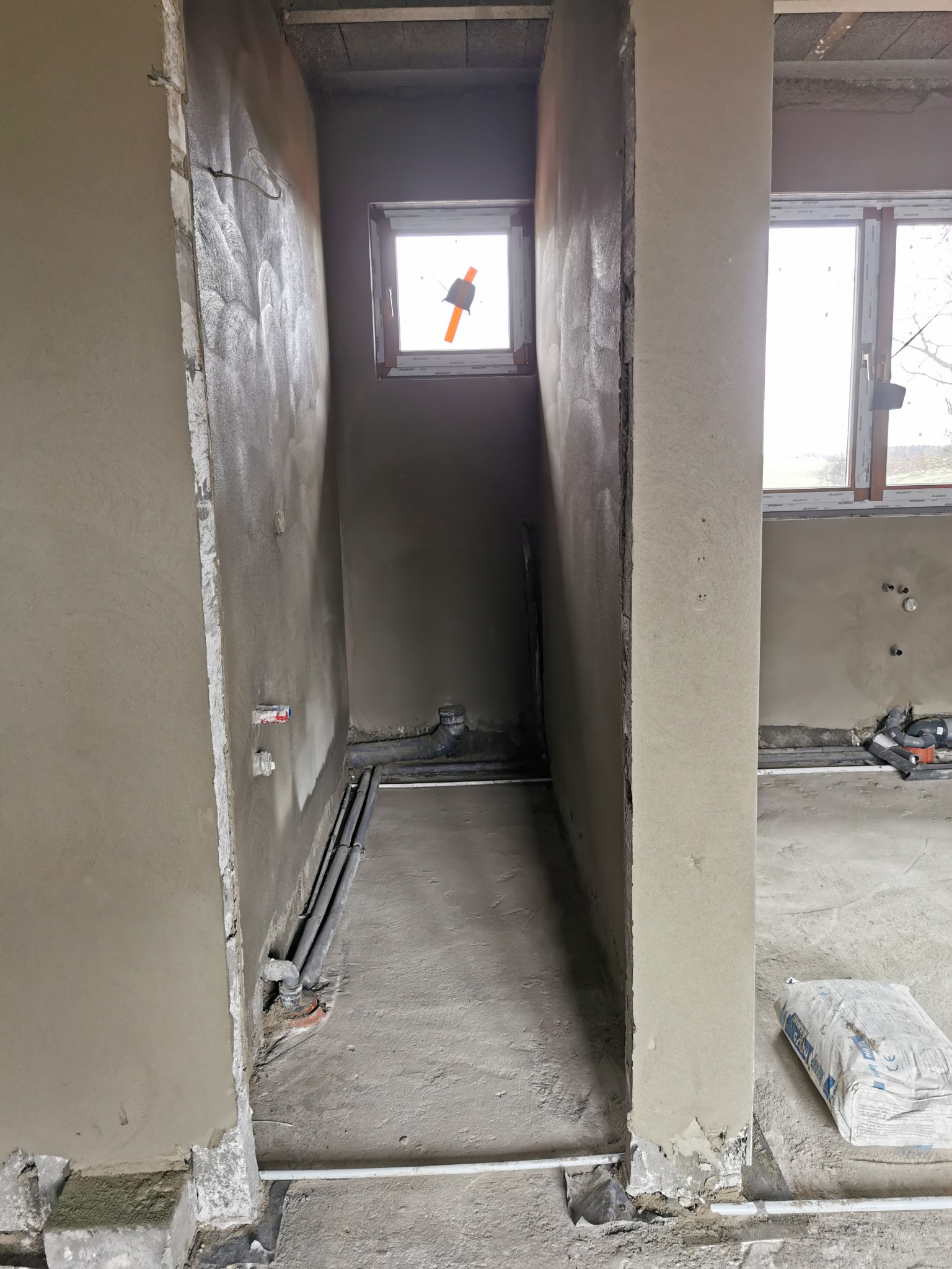 Wnętrze w trakcie prac wykończeniowych: świeżo otynkowane ściany z widocznymi instalacjami, rurami i oknem z zamontowaną folią zabezpieczającą.