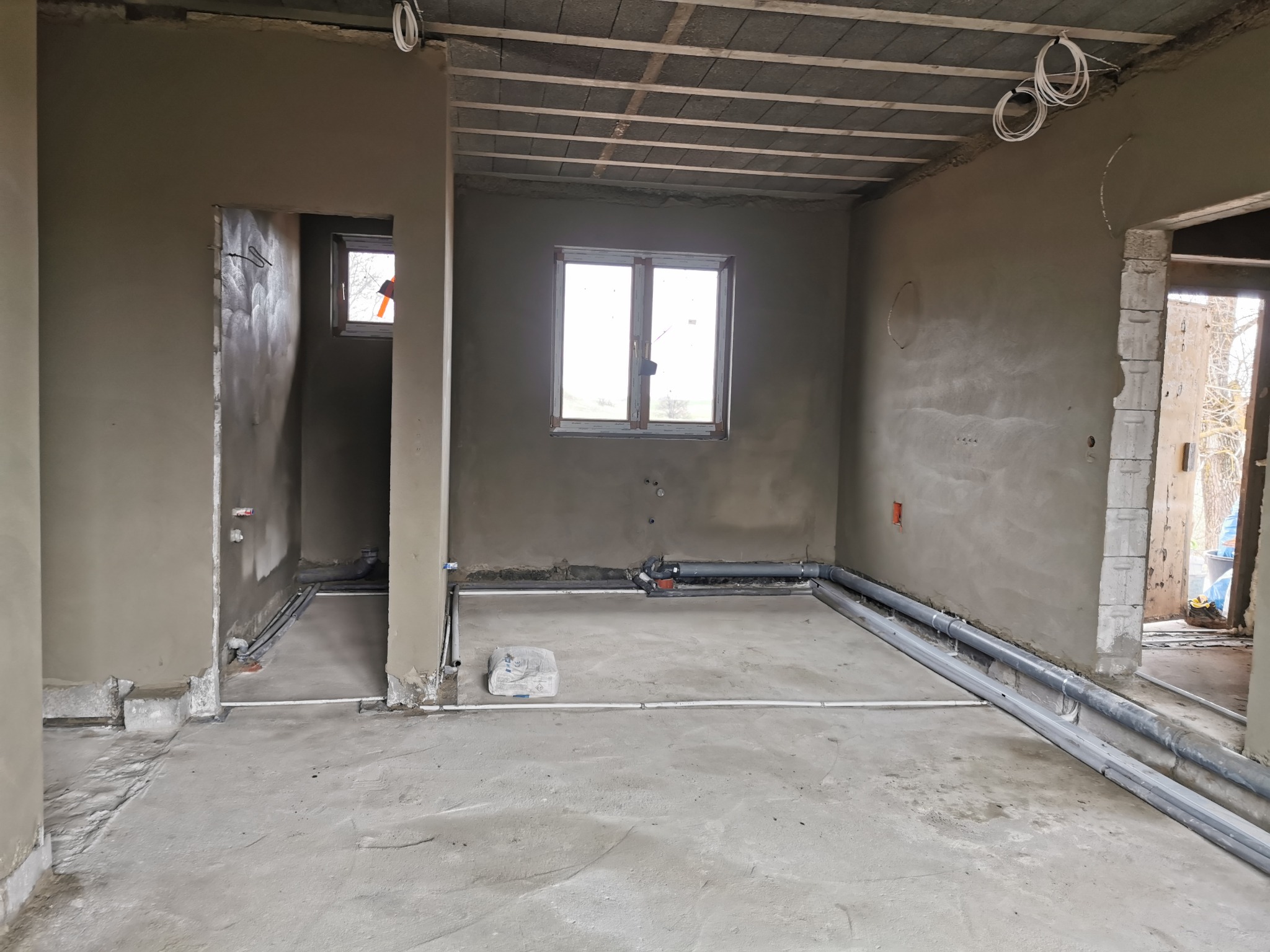 Wnętrze surowego pomieszczenia po nałożeniu szarego tynku maszynowego na ściany, widoczne rury instalacyjne przy podłodze, sufit z betonowych płyt, okno z widokiem na zewnątrz.
