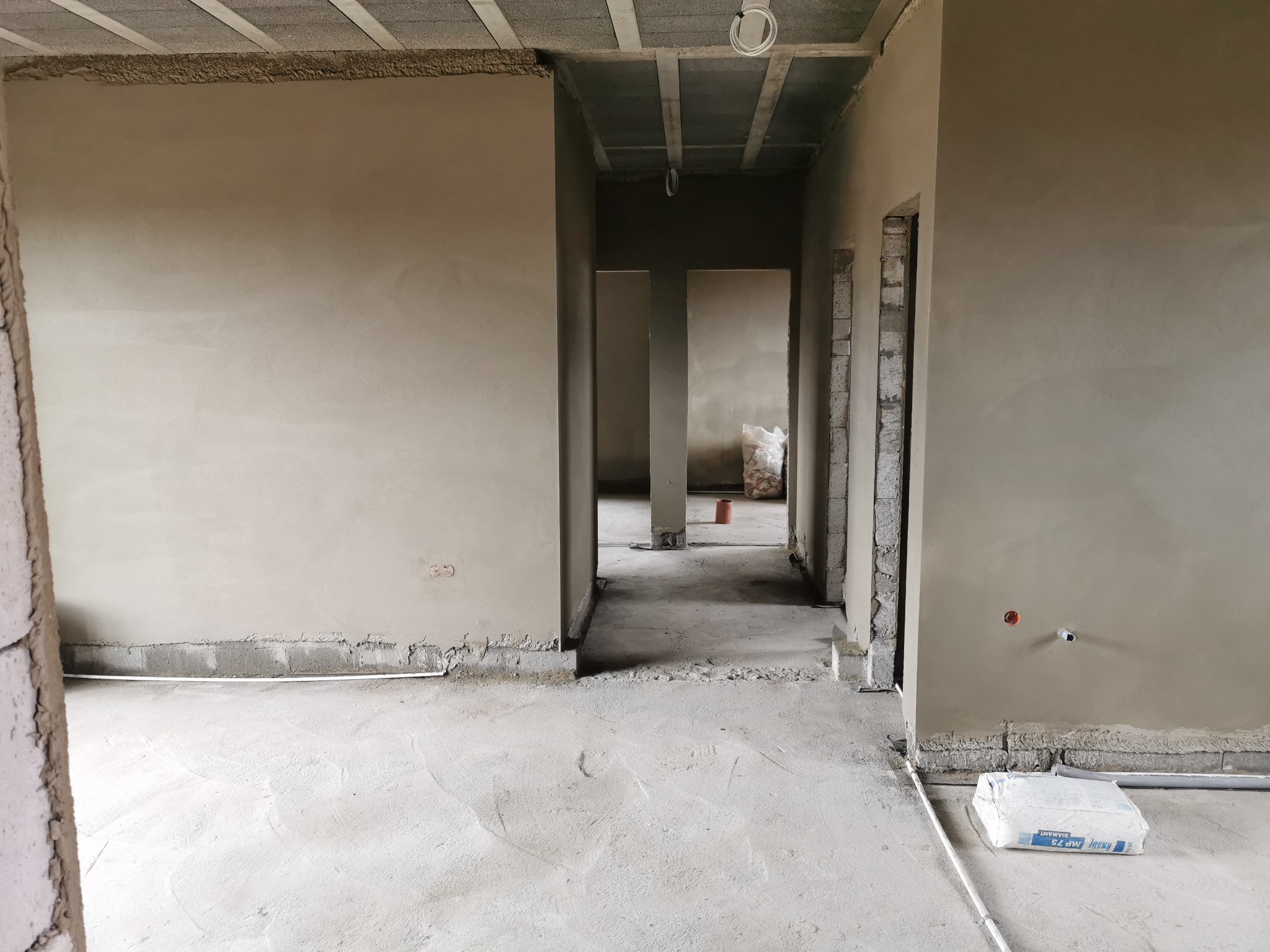 Wnętrze w stanie surowym po nałożeniu gładzi, widoczne ślady prac murarskich przy podłodze, wystające przewody elektryczne i worek z zaprawą na posadzce.
