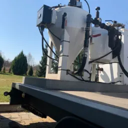 Mobilna instalacja do obróbki strumieniowo-ściernej z dwoma zbiornikami, widoczna na przyczepie w plenerze, z elementami sterującymi i przewodami. Na zbiorniku widoczny napis 'FAN'.