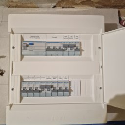 Gerard Gut G-Elektro - Otwarta, biała skrzynka elektryczna Hager z bezpiecznikami, opisana: zmywarka, mikrofala, lodówka, kuchnia, pokój. Widoczne przewody i surowy stan wykończenia ściany.