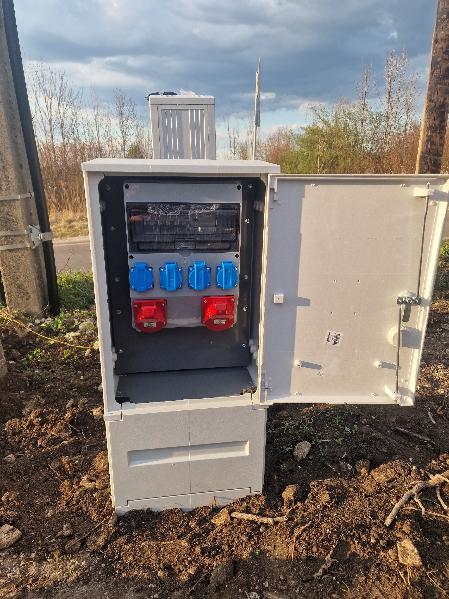 Otwarta skrzynka elektryczna z gniazdami siłowymi (czerwone i niebieskie) na zewnątrz, stojąca na ziemi. Widoczne przewody i elementy instalacji wewnątrz.