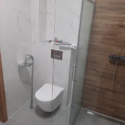 Nowoczesna łazienka z podwieszaną toaletą, kabiną prysznicową typu walk-in z brązowymi płytkami imitującymi drewno i białymi płytkami marmuropodobnymi, widoczne narzędzia i wężyki instalacyjne...