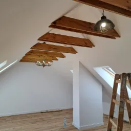 Poddasze po remoncie: białe ściany, drewniane belki stropowe, lampa w stylu industrialnym, drewniana podłoga, drabina malarska i butelka wody mineralnej.