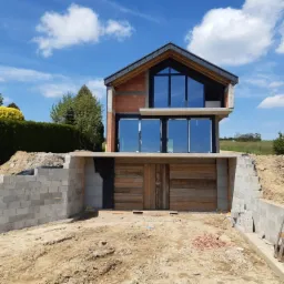 Dwukondygnacyjny dom w trakcie budowy z widoczną surową konstrukcją, dużymi oknami i drewnianą bramą garażową, otoczony placem budowy z blokami betonowymi i ziemią.