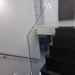 Szklana barierka zamocowana na metalowych uchwytach, czarne schody z podświetleniem LED, metalowa poręcz przy ścianie.