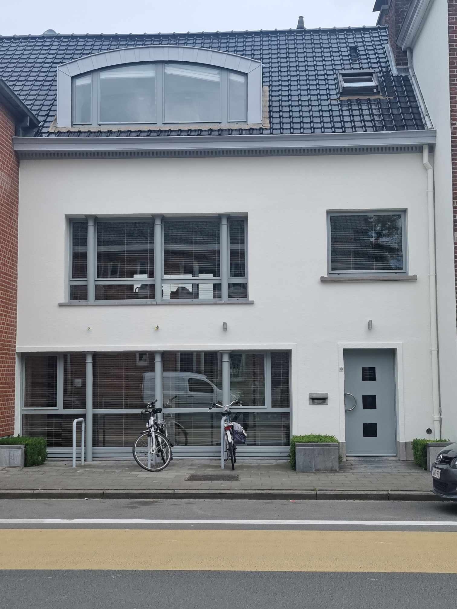 Odnowiona, biała elewacja dwukondygnacyjnego domu z szarymi oknami i drzwiami, widok z ulicy, rowery zaparkowane przed budynkiem.