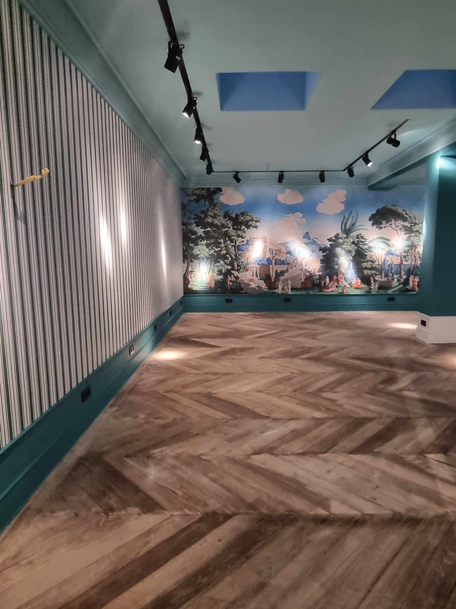 Wnętrze pokoju z parkietem ułożonym w jodełkę, tapetą w pionowe paski i malowidłem na ścianie z motywem krajobrazowym, oświetlone reflektorami na szynoprzewodzie.