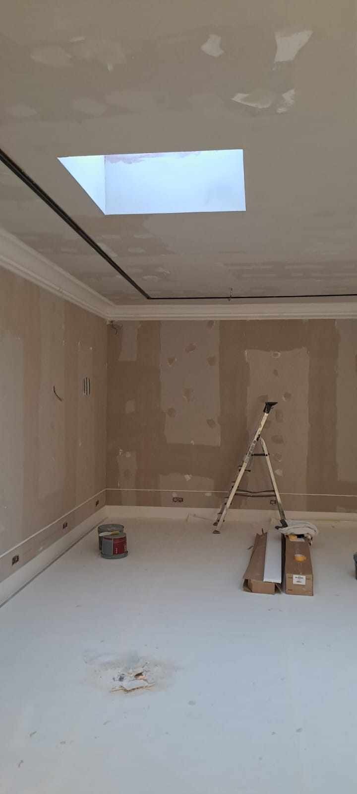 Pokój w trakcie prac wykończeniowych: ściany z widocznymi śladami szpachlowania, sufit z oknem dachowym, drabina, puszki i kartony na podłodze, listwy przypodłogowe.