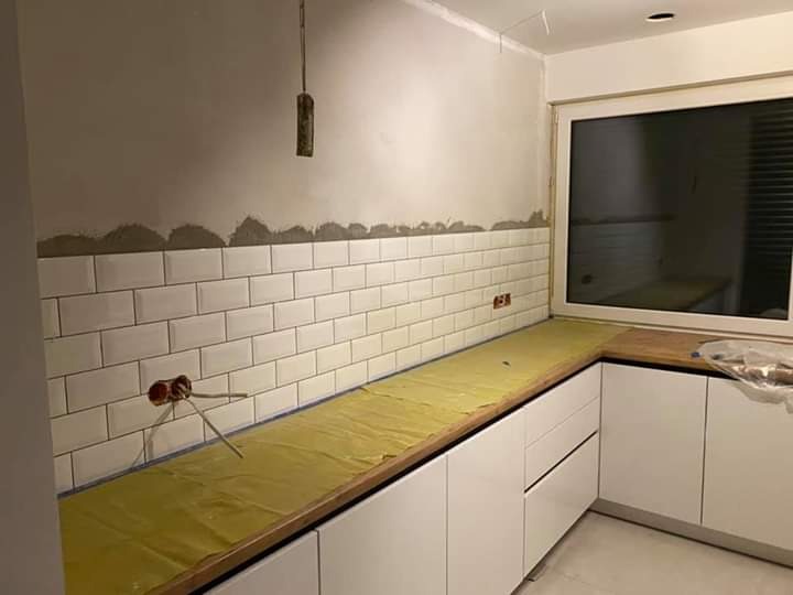 Układanie białych, prostokątnych płytek na ścianie w kuchni, widoczne nieotynkowane fragmenty ściany nad płytkami, zabezpieczony żółtą folią blat i białe szafki kuchenne, wystające kable elektryczne.