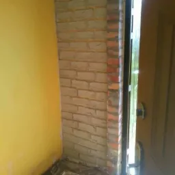 Fragment ściany wewnętrznej wykończonej cegłami klinkierowymi wokół ościeżnicy drzwiowej, widoczna żółta ściana w tle oraz narzędzia budowlane w kuwecie.
