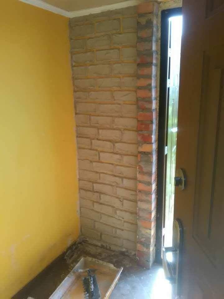 Fragment ściany wewnętrznej wykończonej cegłami klinkierowymi wokół ościeżnicy drzwiowej, widoczna żółta ściana w tle oraz narzędzia budowlane w kuwecie.