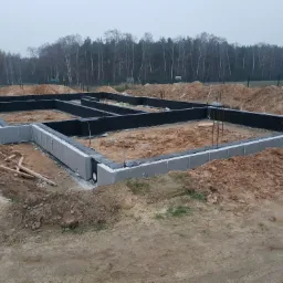 Wylane fundamenty z bloczków betonowych, zabezpieczone czarną izolacją przeciwwilgociową, z wystającymi prętami zbrojeniowymi, na tle piaszczystego terenu i drzew.
