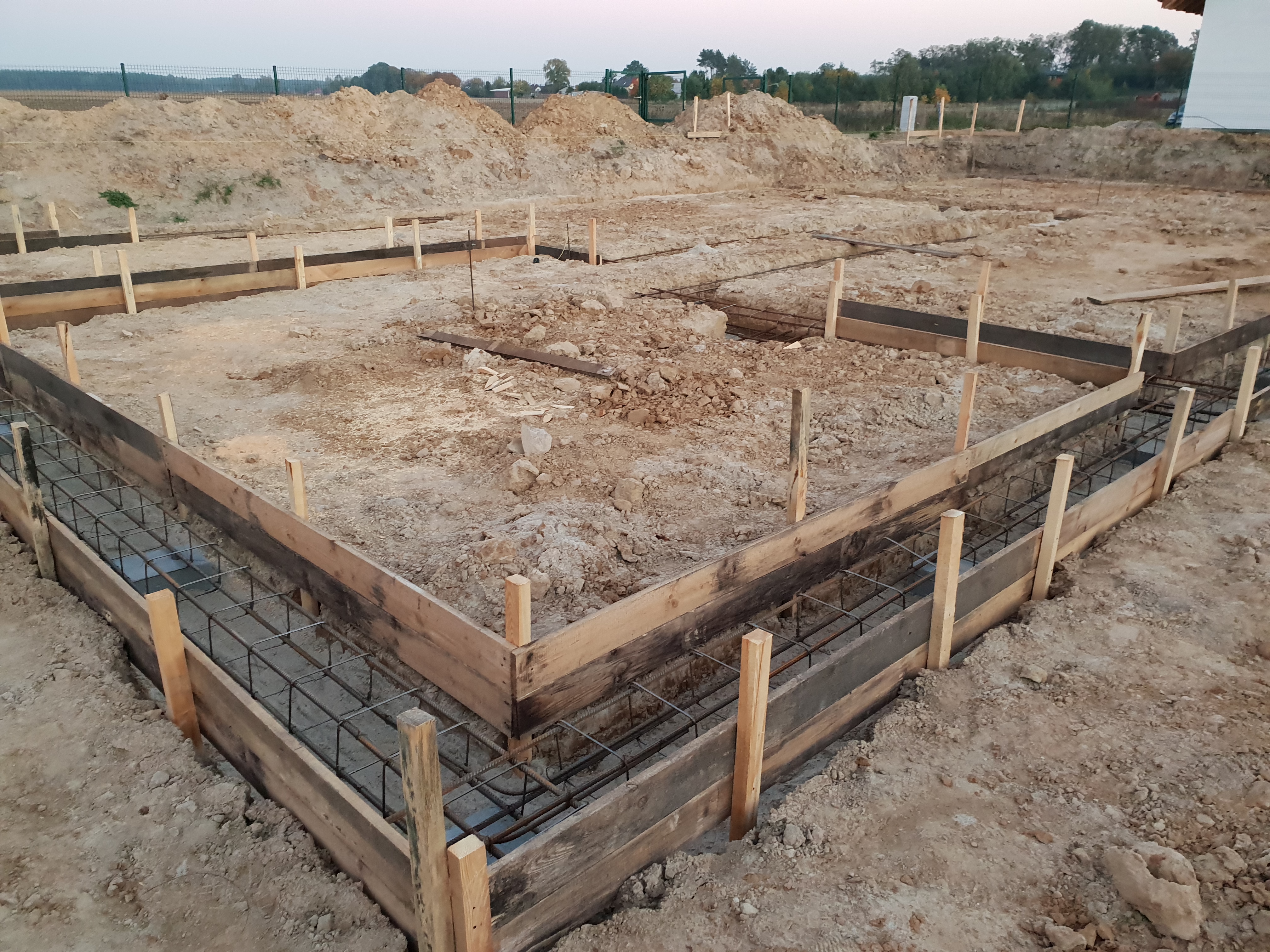 Zbrojenie fundamentów w drewnianym szalunku, widok na wykop z ziemią i ogrodzeniem w tle.
