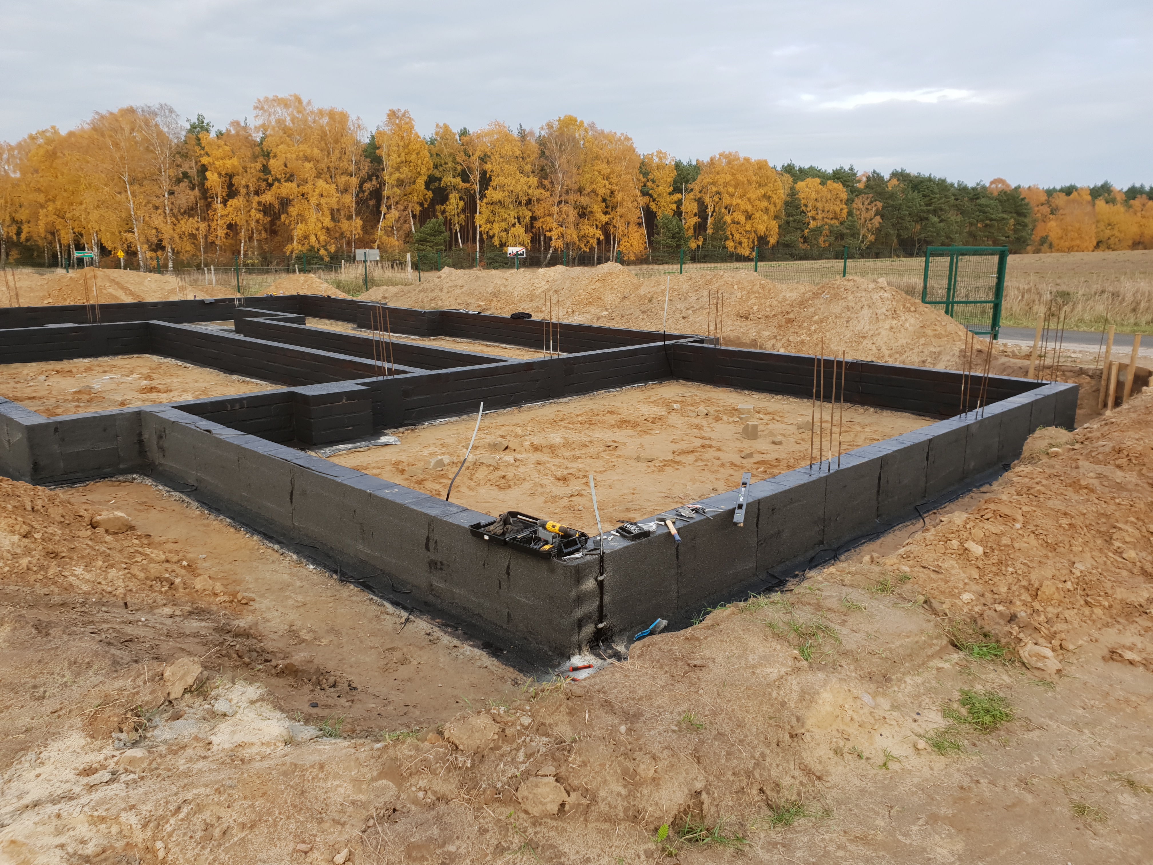 Zalane fundamenty z wystającymi prętami zbrojeniowymi, ocieplone czarnym styropianem, na tle jesiennego krajobrazu z drzewami o żółtych liściach i pochmurnym niebem.