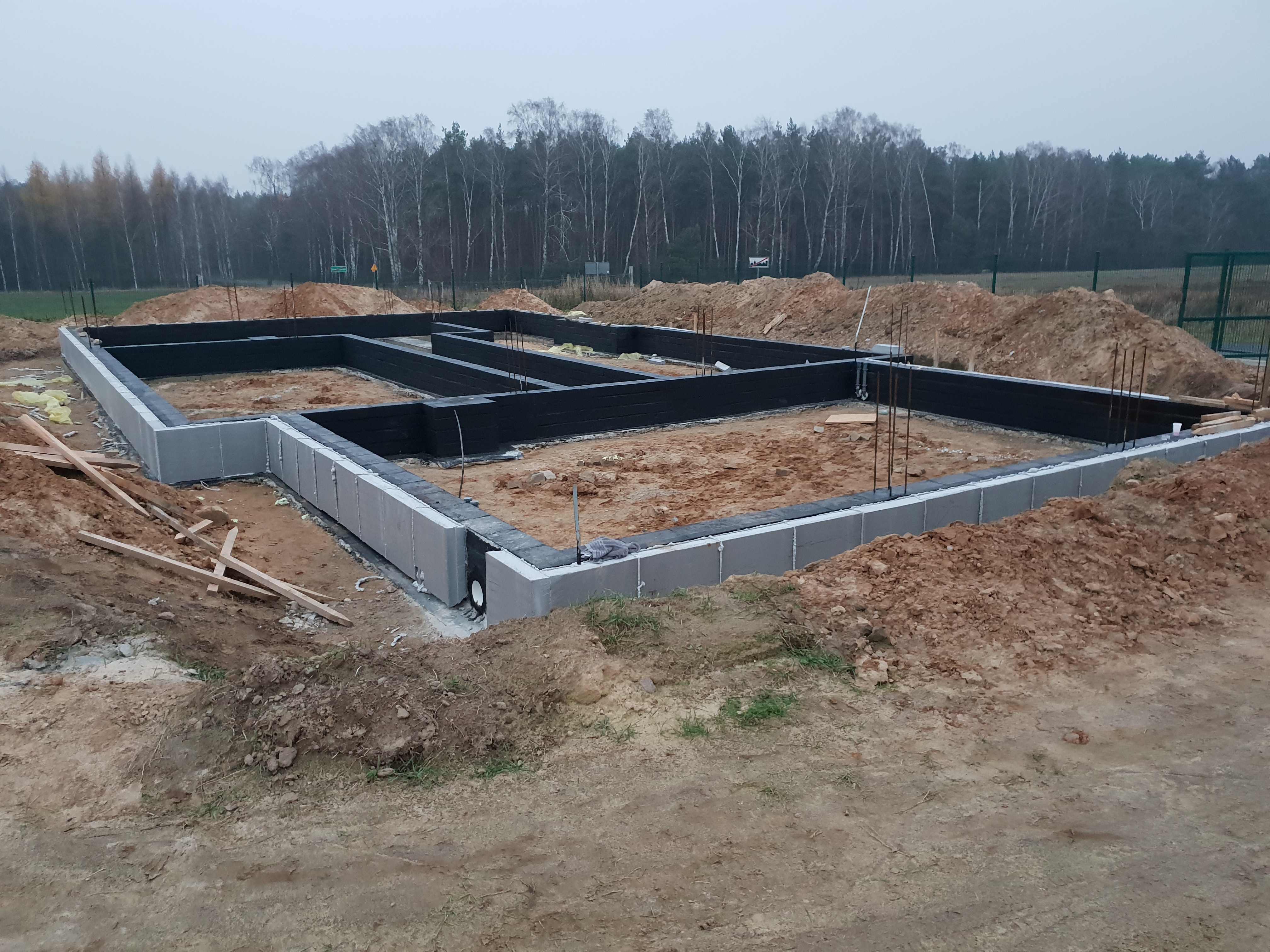 Wylane fundamenty z bloczków betonowych, zabezpieczone czarną izolacją przeciwwilgociową, z wystającymi prętami zbrojeniowymi, na tle piaszczystego terenu i drzew.