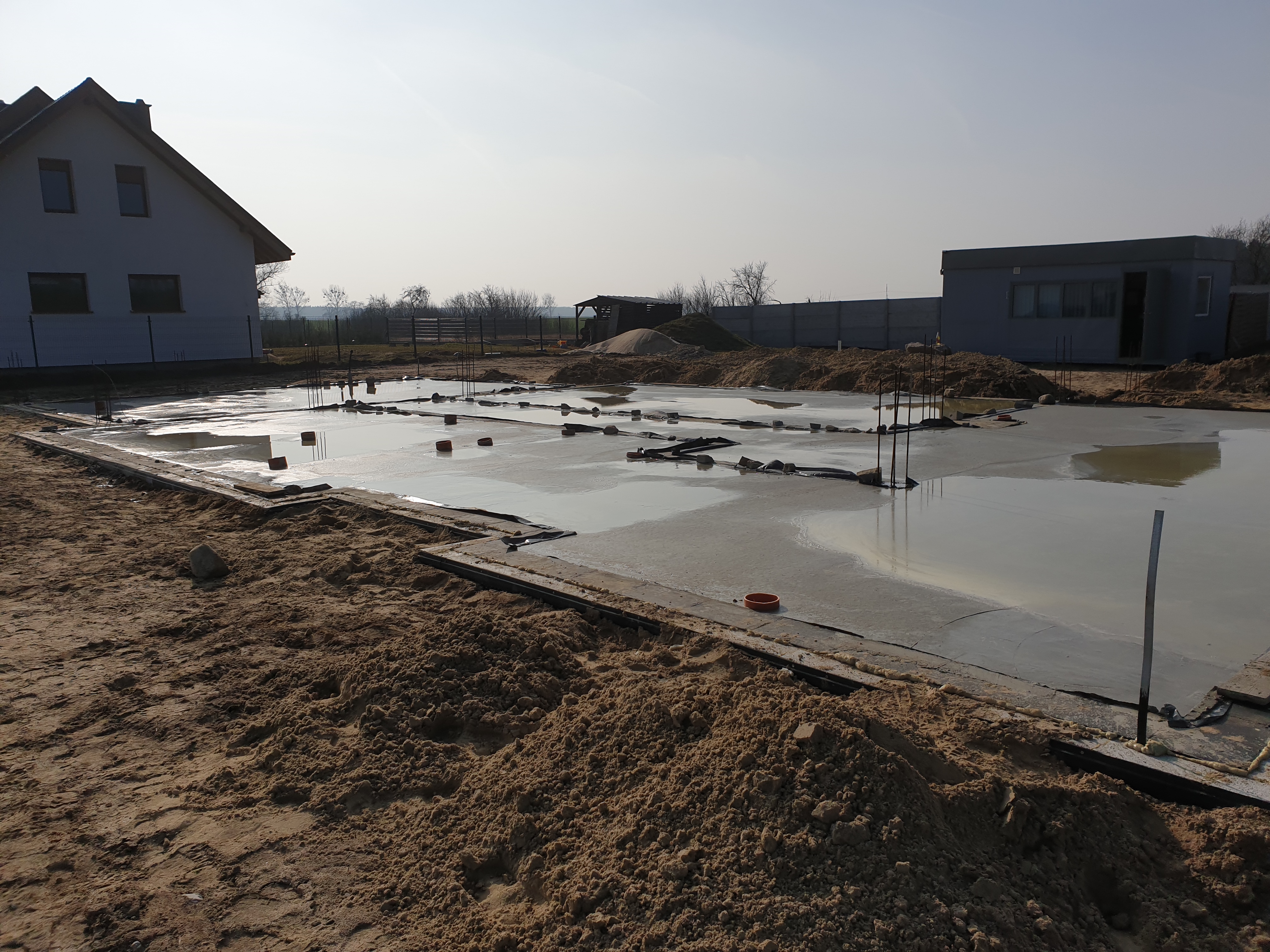Wylany beton na przygotowany szalunek fundamentów budynku jednorodzinnego, widoczne wystające pręty zbrojeniowe i elementy izolacji przeciwwilgociowej.