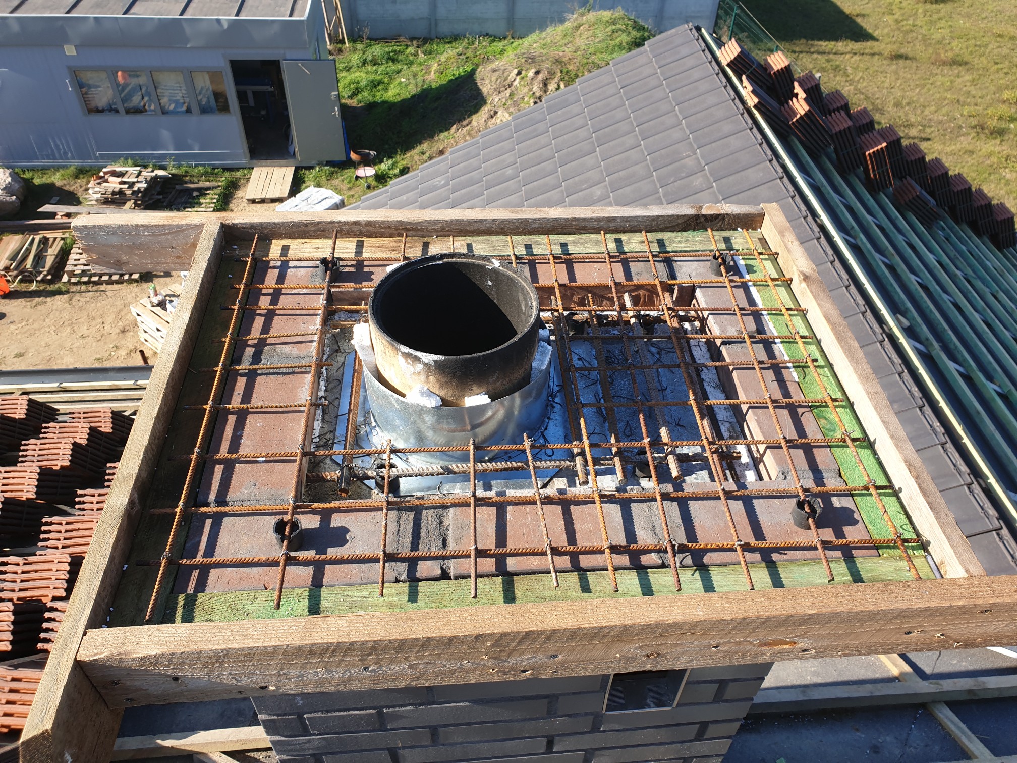 Konstrukcja zbrojeniowa wokół komina na dachu w trakcie budowy, widoczne cegły, drewniane elementy szalunku oraz ułożone dachówki na sąsiedniej połaci dachu.