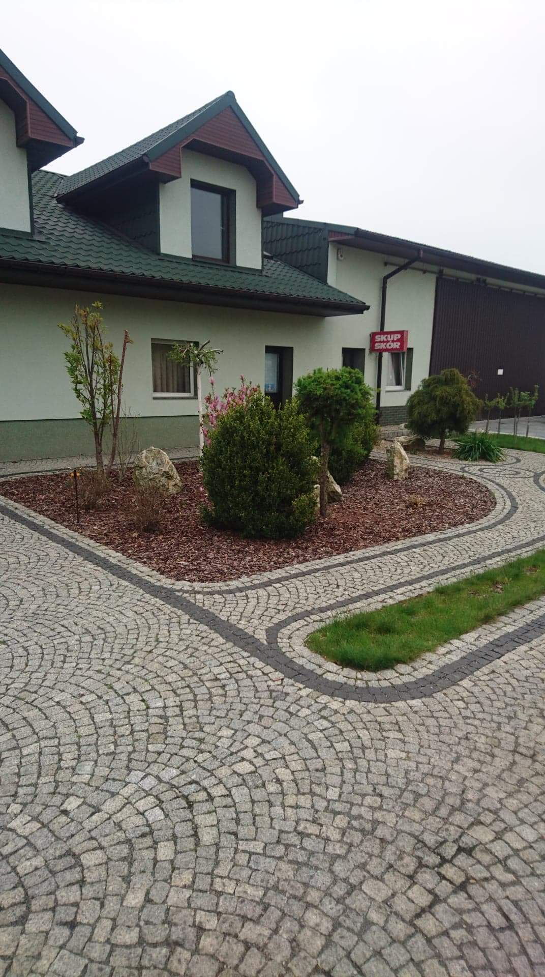 Granitowa ścieżka ułożona w łukowaty wzór, otaczająca rabatę z krzewami i kamieniami, na tle budynku ze znakiem 'Skup Skór'.