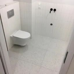 Nowoczesna łazienka wykończona w bieli z akcentami czerni, z wiszącą toaletą, armaturą i płytkami imitującymi lastryko na podłodze.