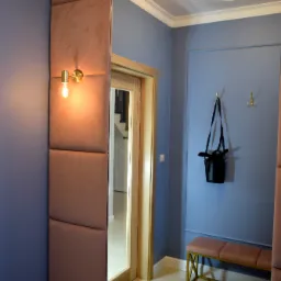 Przedpokój w odcieniach błękitu i pudrowego różu z dużym lustrem w ramie z paneli tapicerowanych, złotym kinkietem i minimalistyczną ławeczką na złotym stelażu, widok na schody przez otwarte drzwi.