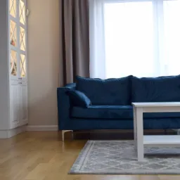 Niebieska sofa welurowa z poduszkami, biały stolik kawowy z kwiatami w wazonie, szary dywan ze wzorem, biała szafka z oświetleniem w tle, ściana w kolorze granatowym.